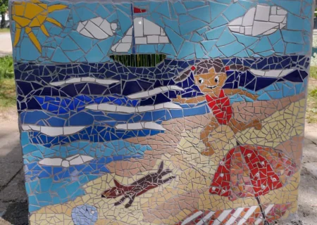 Ein farbenfrohes Mosaik zeigt ein Kind, das am Strand mit einem Hund läuft, mit einem Segelboot im Hintergrund. Angelika Schaupps Beitrag für das internationale Projekt, symbolisiert den Sommer.