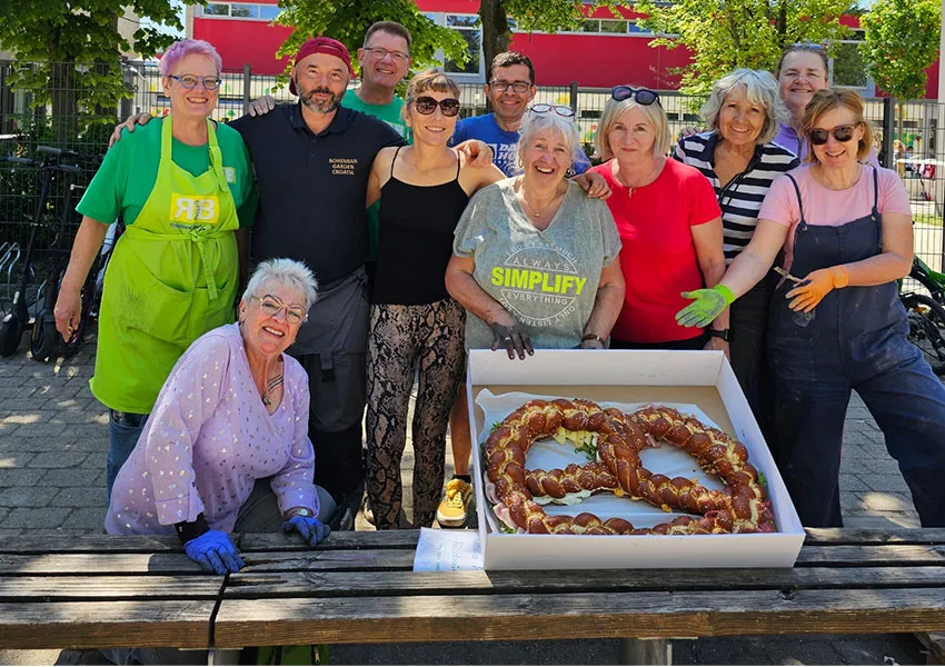 Angelika Schaupp vom Pflegedienst AML posiert mit einer großen Brezel, um ihren Beitrag zum internationalen Mosaikprojekt in Ingolstadt zu feiern. Das Mosaik symbolisiert die vier Jahreszeiten.