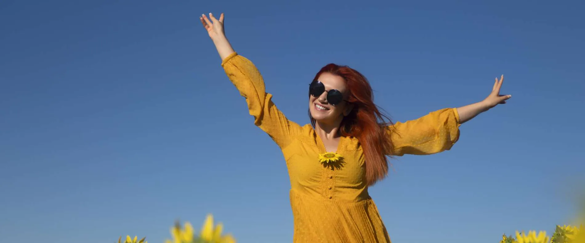 Eine Frau in gelbem Kleid und Sonnenbrille streckt die Arme freudig in die Luft unter einem klaren blauen Himmel, symbolisiert die positive Energie und die neuen Perspektiven, die ein Quereinstieg in die Pflege bietet.