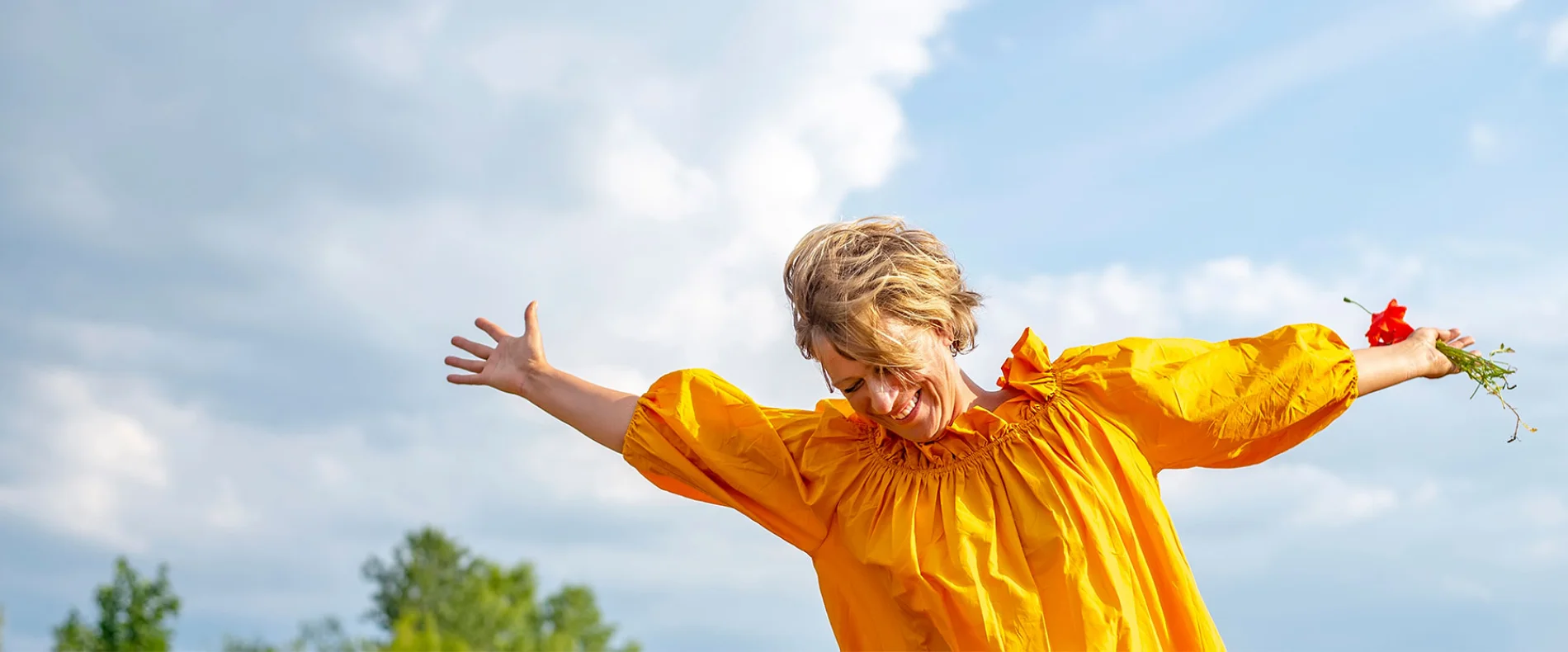 Freudestrahlende Frau in gelbem Kleid streckt die Arme in einem sonnigen Feld aus, symbolisiert Lebensfreude und Freiheit.