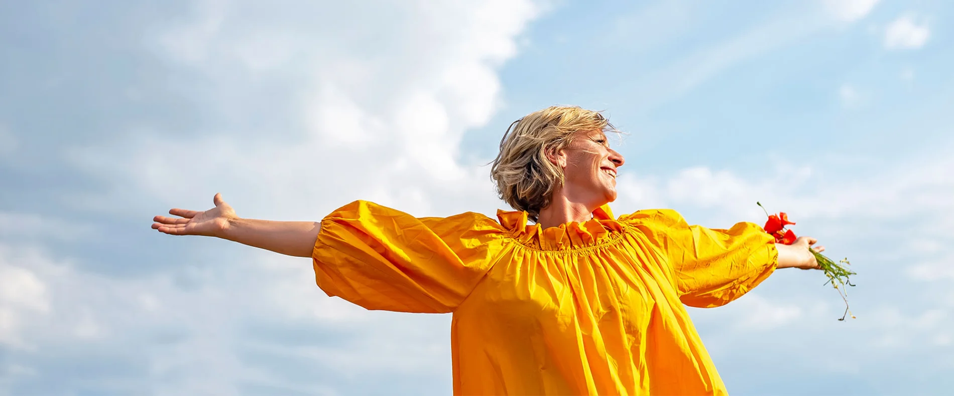 Eine Frau in einer gelben Bluse steht mit ausgebreiteten Armen vor einem blauen Himmel und lächelt. Dieses Bild symbolisiert die Freude und Leidenschaft, die Pflegekräfte bei Pflegedienst AML in Röhrmoos ausstrahlen.